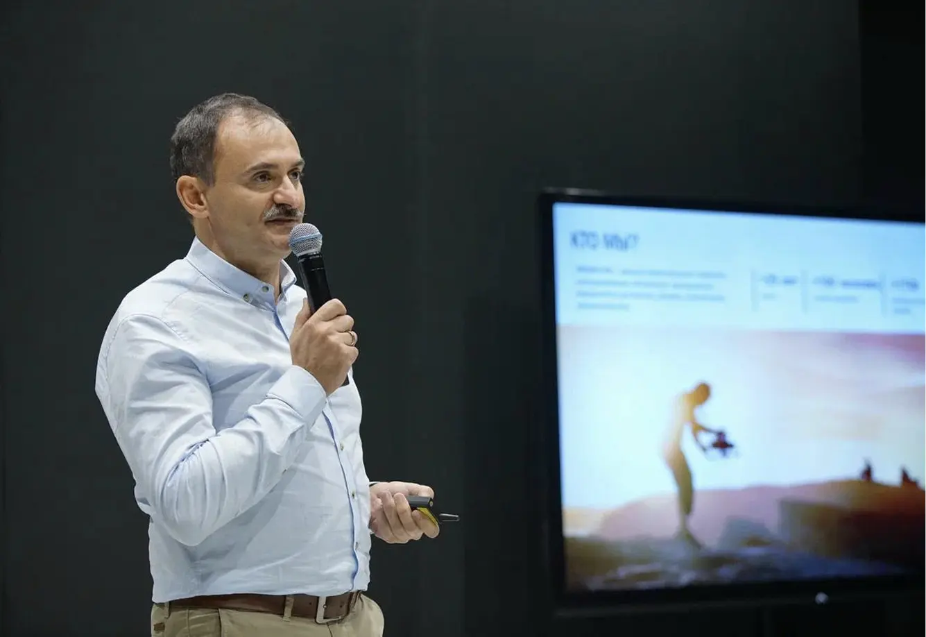 Мужчина в светлой рубашке держит микрофон и выступает перед аудиторией, рядом — большой экран с презентационным слайдом, на котором изображён человек с дроном на фоне заката.