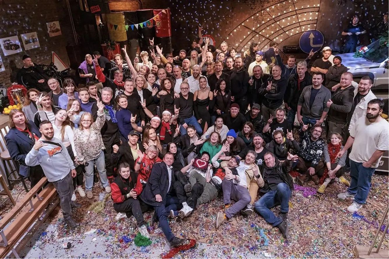Большая группа людей позирует для общего фото на праздничной площадке, усыпленной конфетти; многие улыбаются, машут руками и показывают жесты «peace», вокруг — яркие декорации и праздничная атмосфера.
